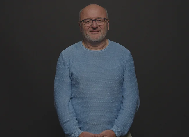 A man wearing a blue sweatshirt smiles at the camera and holds his arms loosely in front of him.
