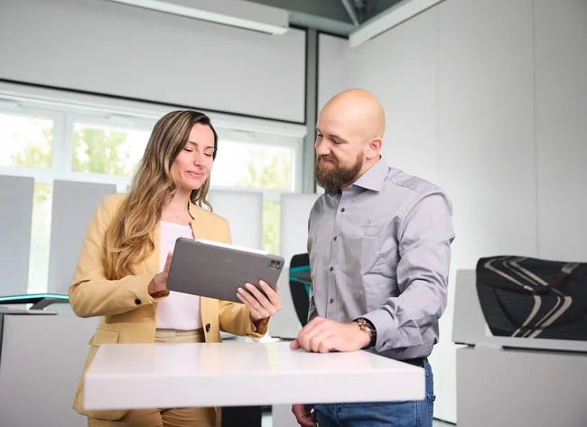 A man and a woman are standing at a table. The woman is showing the man something on a screen.