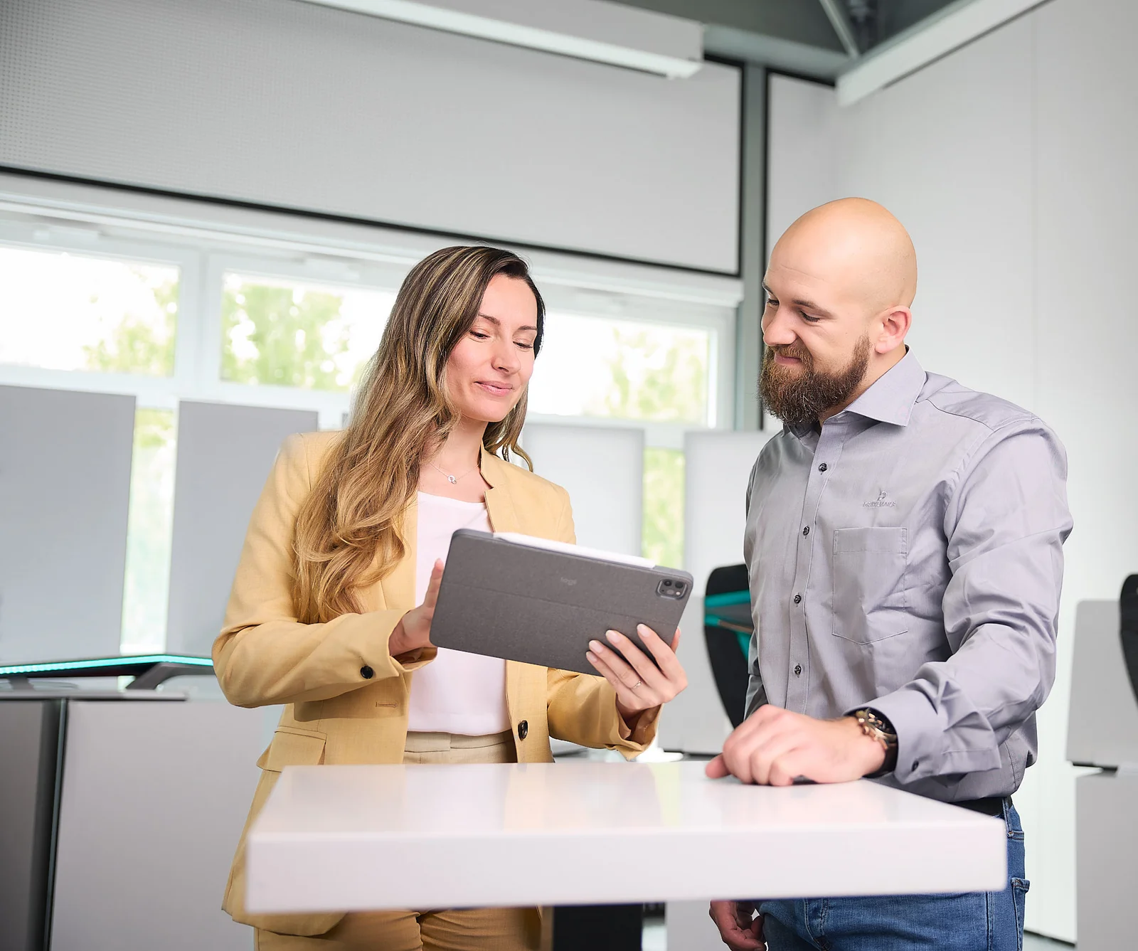 A man and a woman are standing at a table. The woman is showing the man something on a screen.