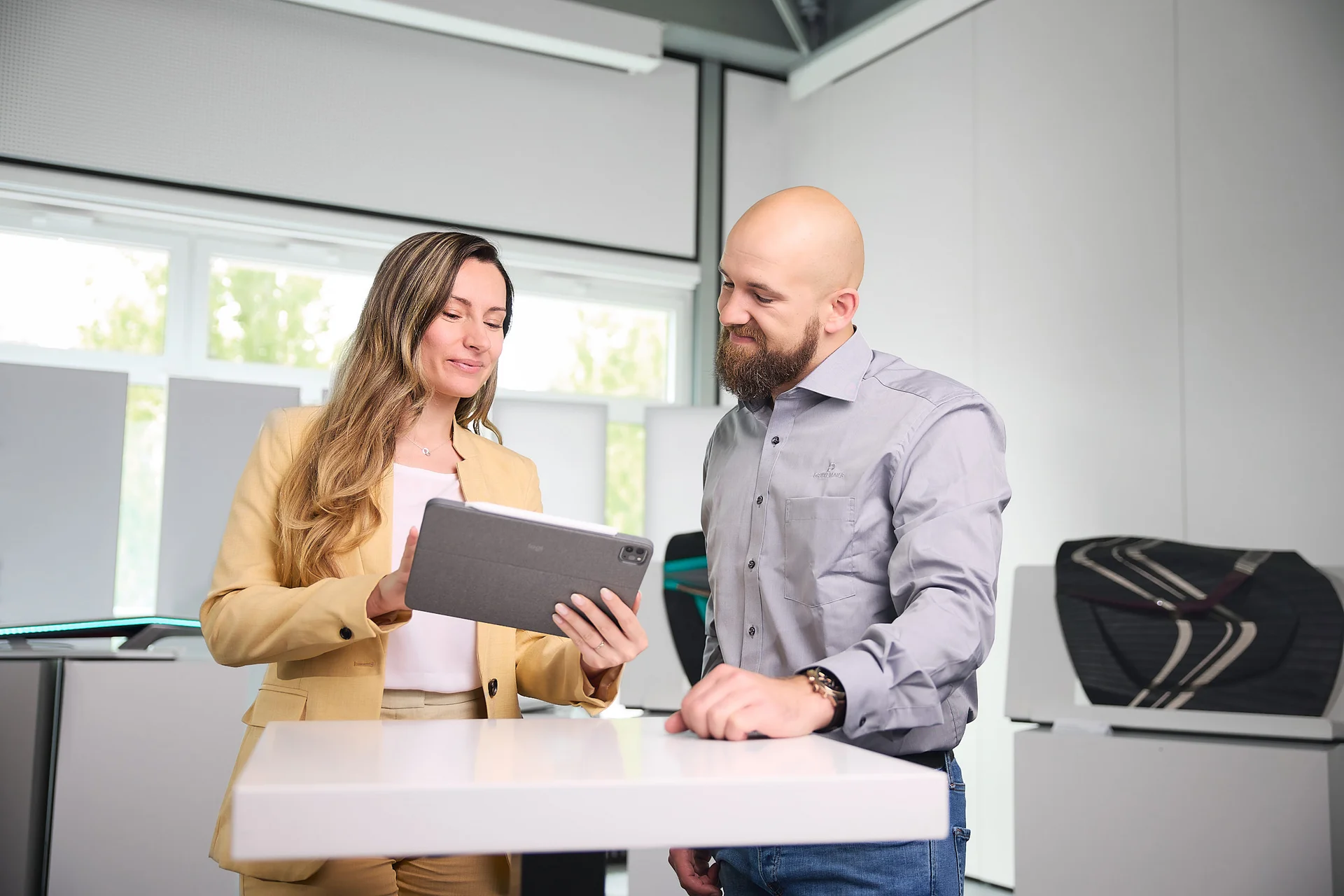 A man and a woman are standing at a table. The woman is showing the man something on a screen.