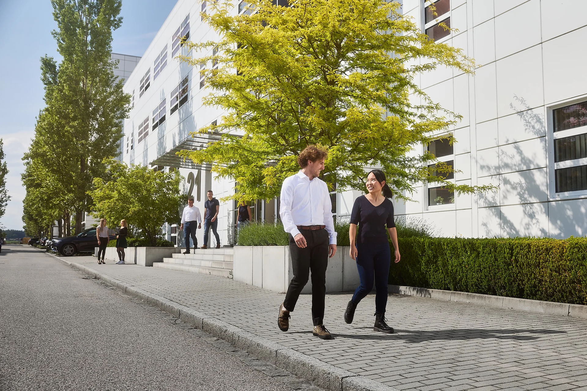 Two people are walking along a path and talking. In the background is a DRÄXLMAIER building and trees.