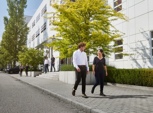 Two people are walking along a path and talking. In the background is a DRÄXLMAIER building and trees.