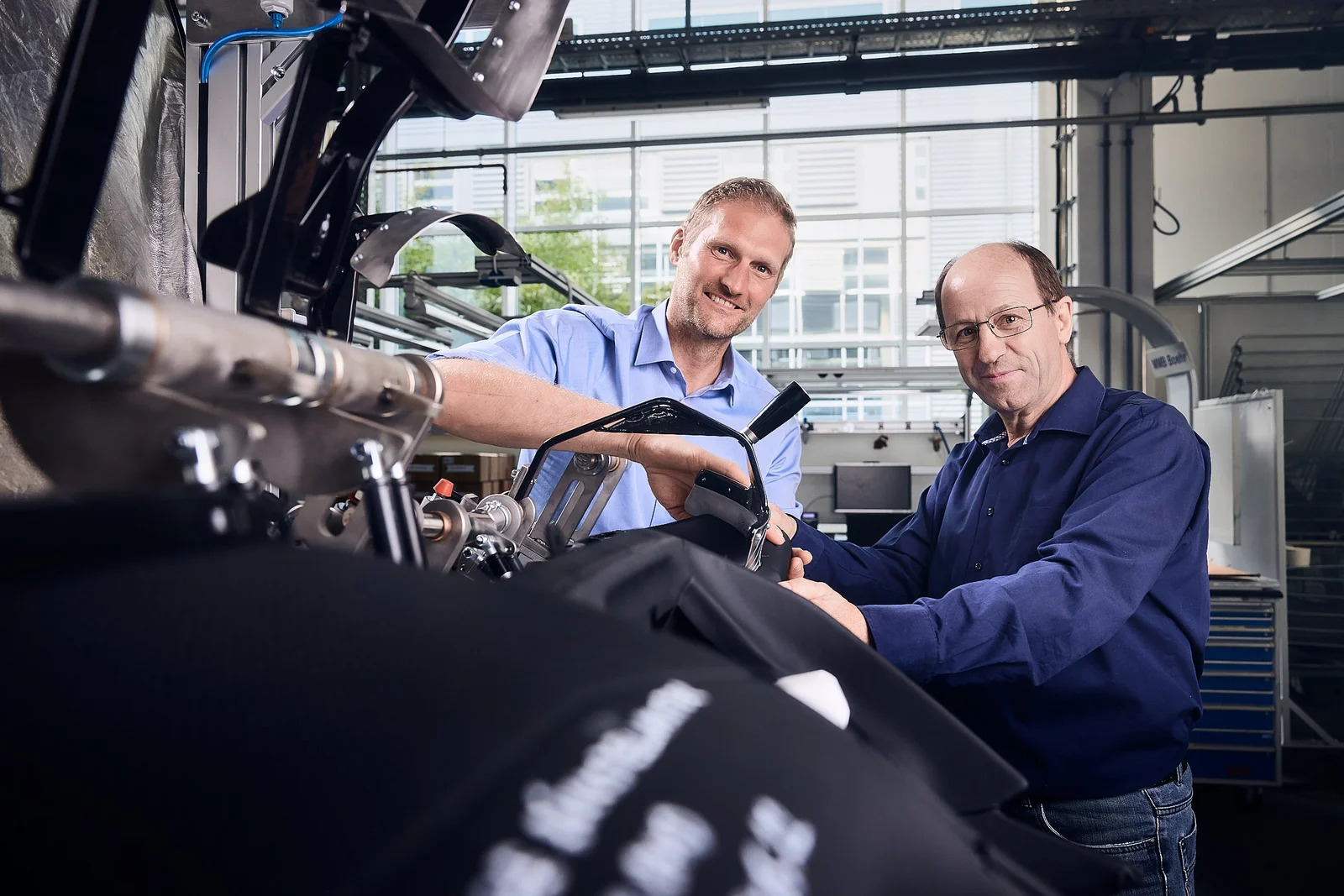 Zwei Mitarbeiter lächeln in die Kamera. Vor ihnen befindet sich ein noch nicht fertiggestellter Fahrzeuginnenraum.