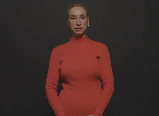 A woman in an orange dress holds her arms loosely in front of her body and looks at the camera.
