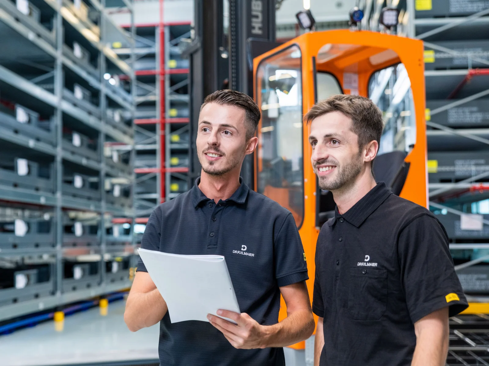 Zwei Lagerlogistiker im Werk Sachsenheim. Im Hintergrund ein orangener Hochhubwagen und das große Lager.
