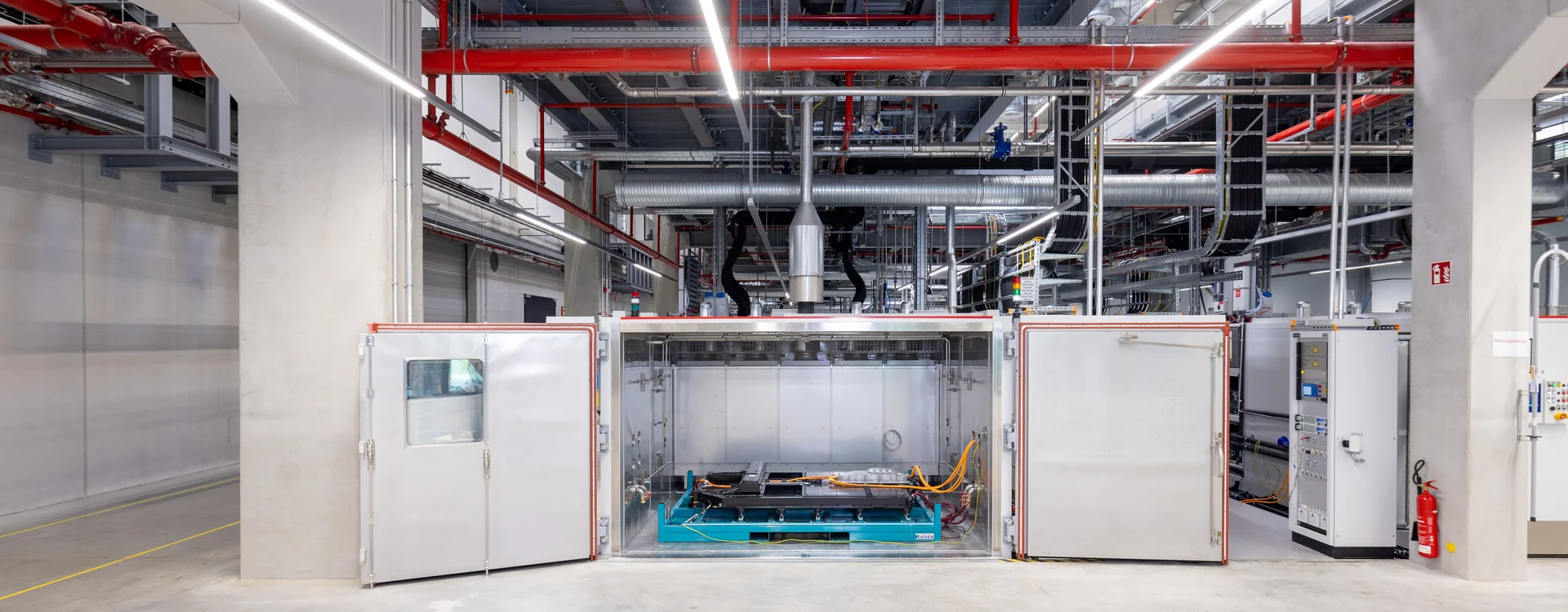 Industrial environment with a large machine in the center and various pipes and cables on the ceiling.