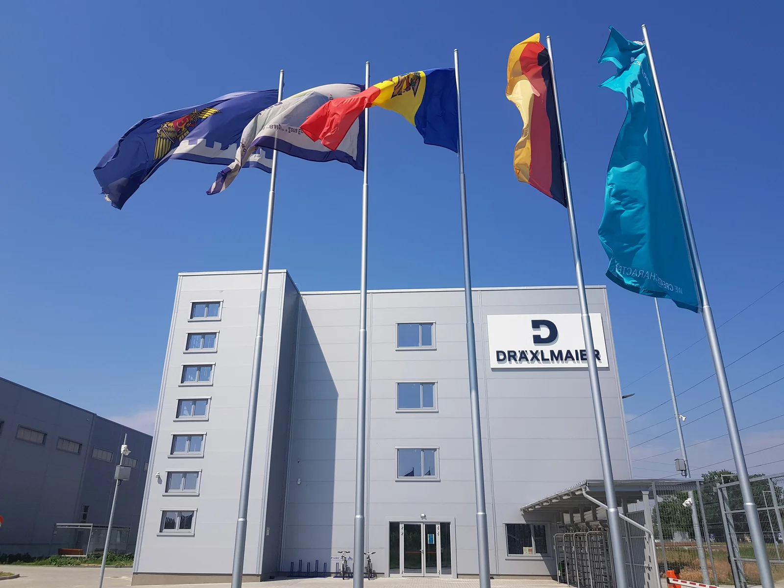 Fünf Fahnen vor einem modernen Gebäude in Cahul mit dem Logo von DRÄXLMAIER und blauem Himmel im Hintergrund.
