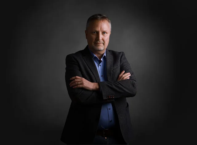 A man stands smiling with his arms folded in front of a black background.