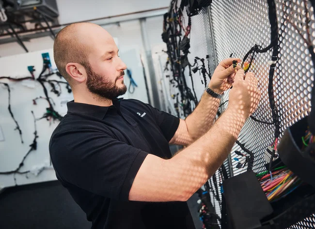A man in a black polo shirt is working on a wiring harness attached to a perforated metal wall.