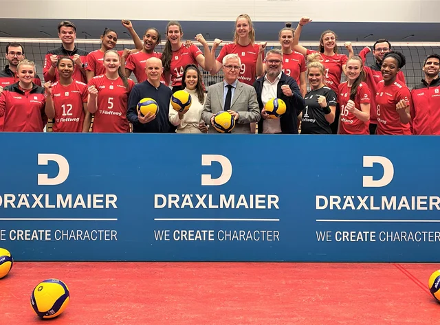 DRÄXLMAIER employees cheer with the Rote Raben volleyball players behind a DRÄXLMAIER banner.