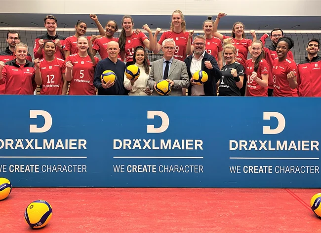 Mitarbeiter von DRÄXLMAIER stehen mit den Volleyballspielerinnen der Roten Raben jubelnd hinter einem DRÄXLMAIER-Banner.