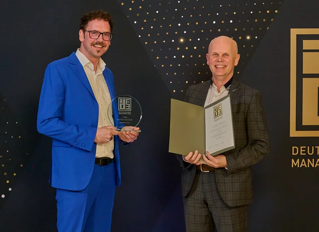 Two men in suits in front of a black wall. The German Ideas Management Award is being presented. 