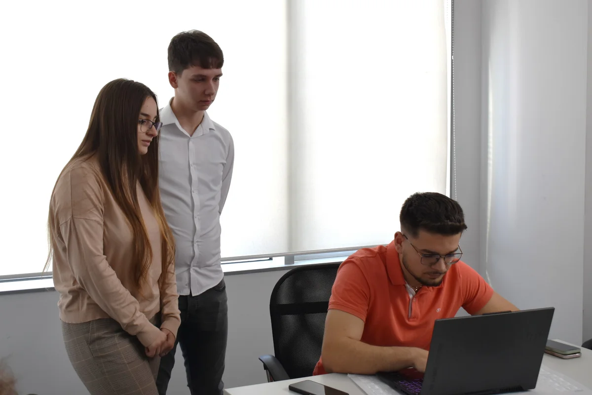 Drei Personen in einem Büro, eine sitzt am Laptop, zwei stehen