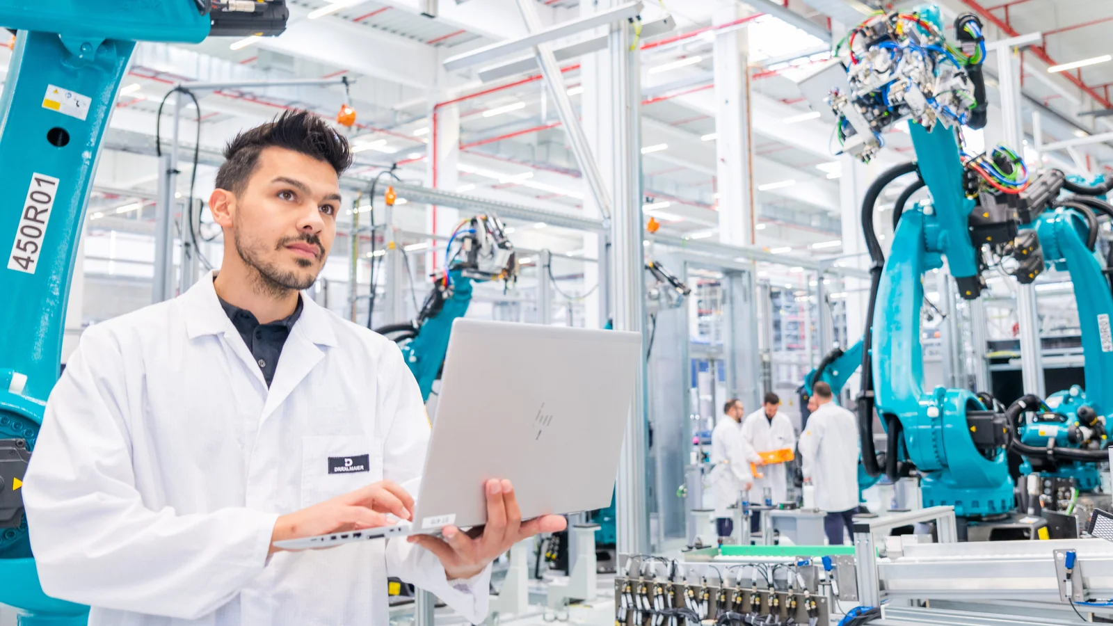 Ein Mann in einem weißen Laborkittel arbeitet an einem Laptop in einer Fabrikhalle mit mehreren blauen Industrierobotern.
