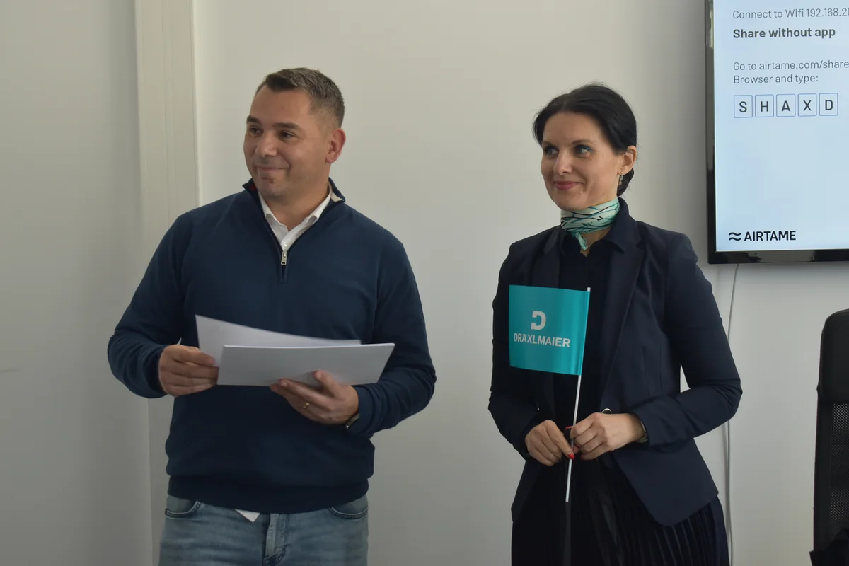 Zwei Personen mit Papier und kleiner Fahne vor Präsentationsfolie mit Gliederungspunkten.