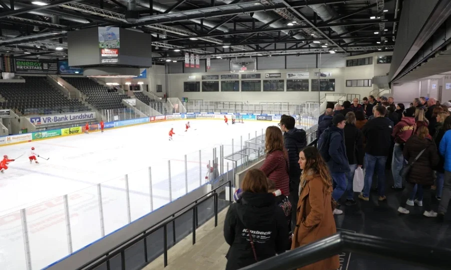 Eishalle mit Spielern auf dem Eis und Zuschauern, die entlang eines Geländers stehen und das Spiel beobachten. Ein großer Bildschirm hängt von der Decke über der Eisfläche.
