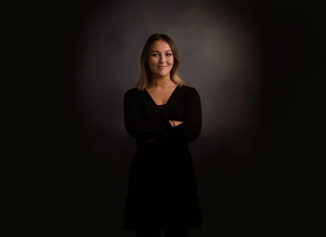 A woman in front of a black background smiles into the camera with her arms crossed. 