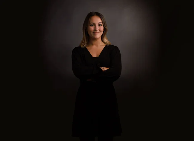 A woman in front of a black background smiles into the camera with her arms crossed. 