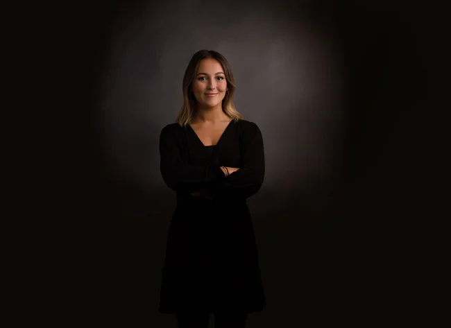 A woman in front of a black background smiles into the camera with her arms crossed. 