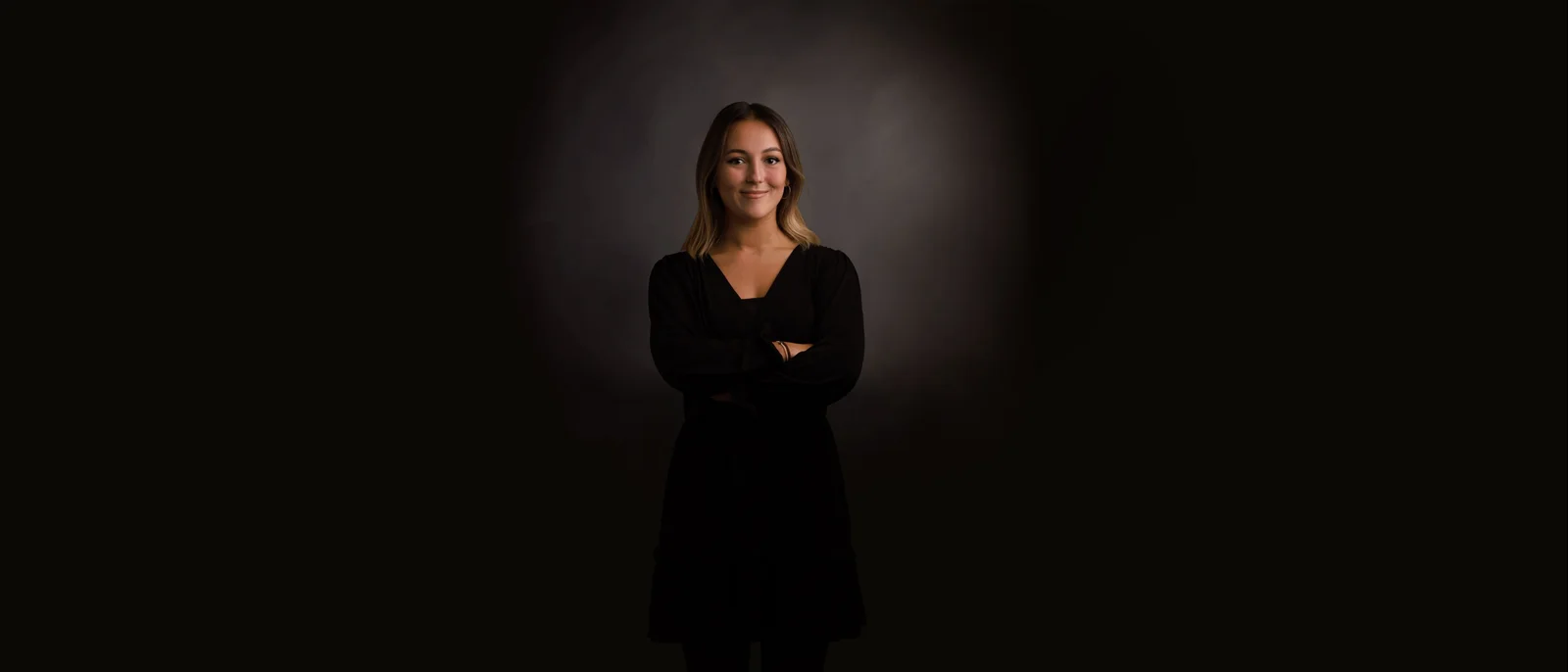 A woman in front of a black background smiles into the camera with her arms crossed. 
