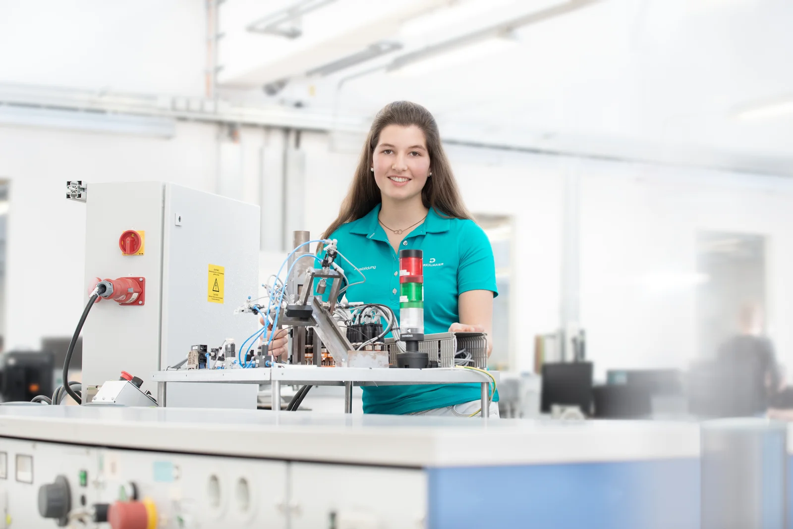 An electronics apprentice at DRÄXLMAIER in Vilsbiburg smiles at the camera and works on a circuit.