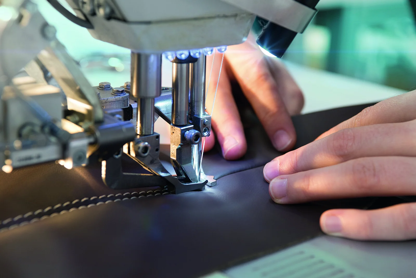 A sewing machine sews a seam on a piece of brown leather, while two hands guide the material.