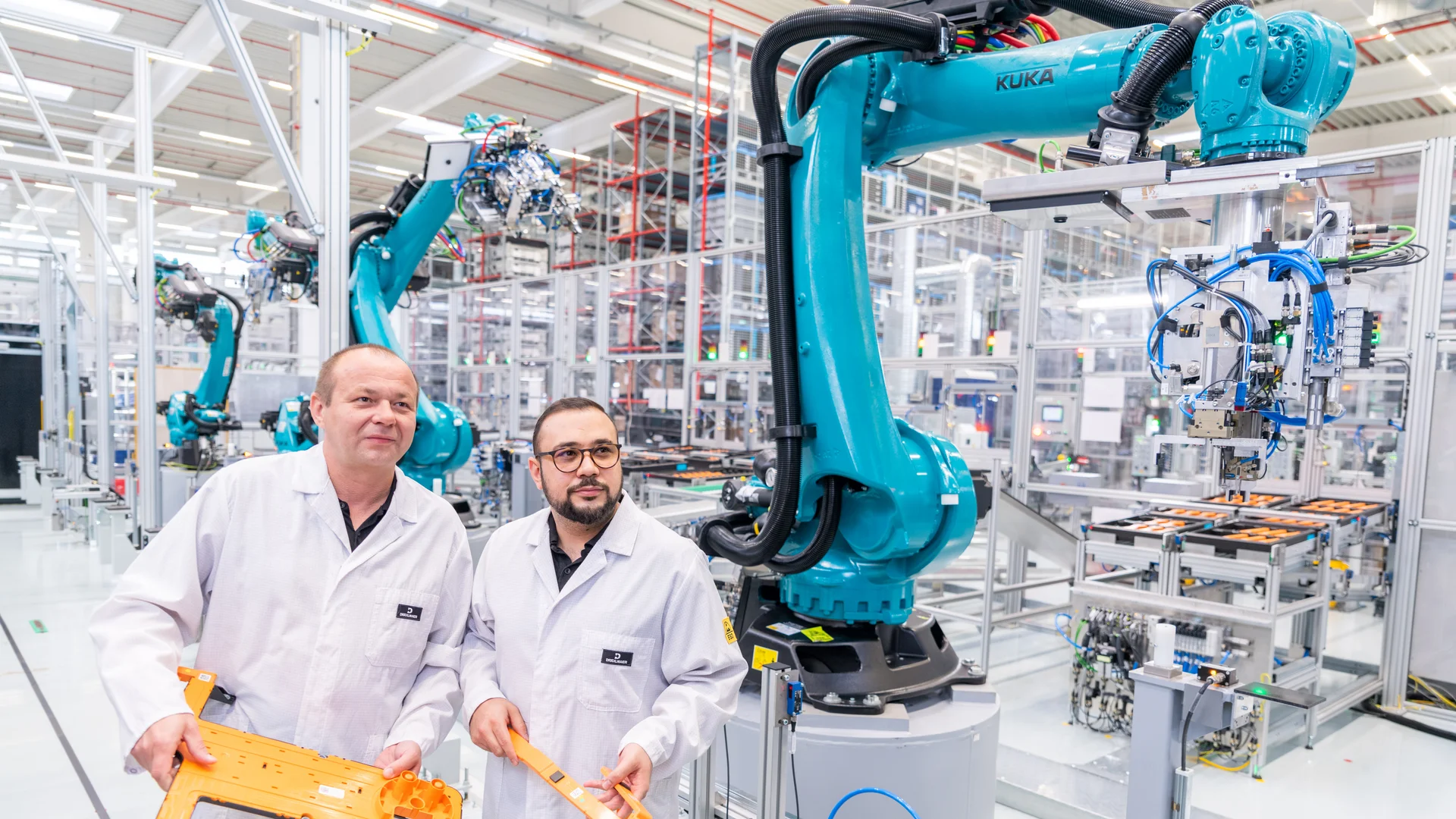 Zwei DRÄXLMAIER Produktionsmitarbeiter in weißem Kittel in einer Produktionshalle. 