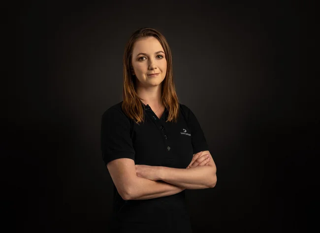A woman in front of a black background smiles at the camera with her arms crossed.