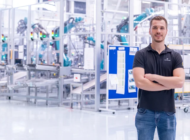 Mann lächelt mit verschränkten Armen in die Kamera. Im Hintergrund sind Produktionsanlagen zu sehen. 