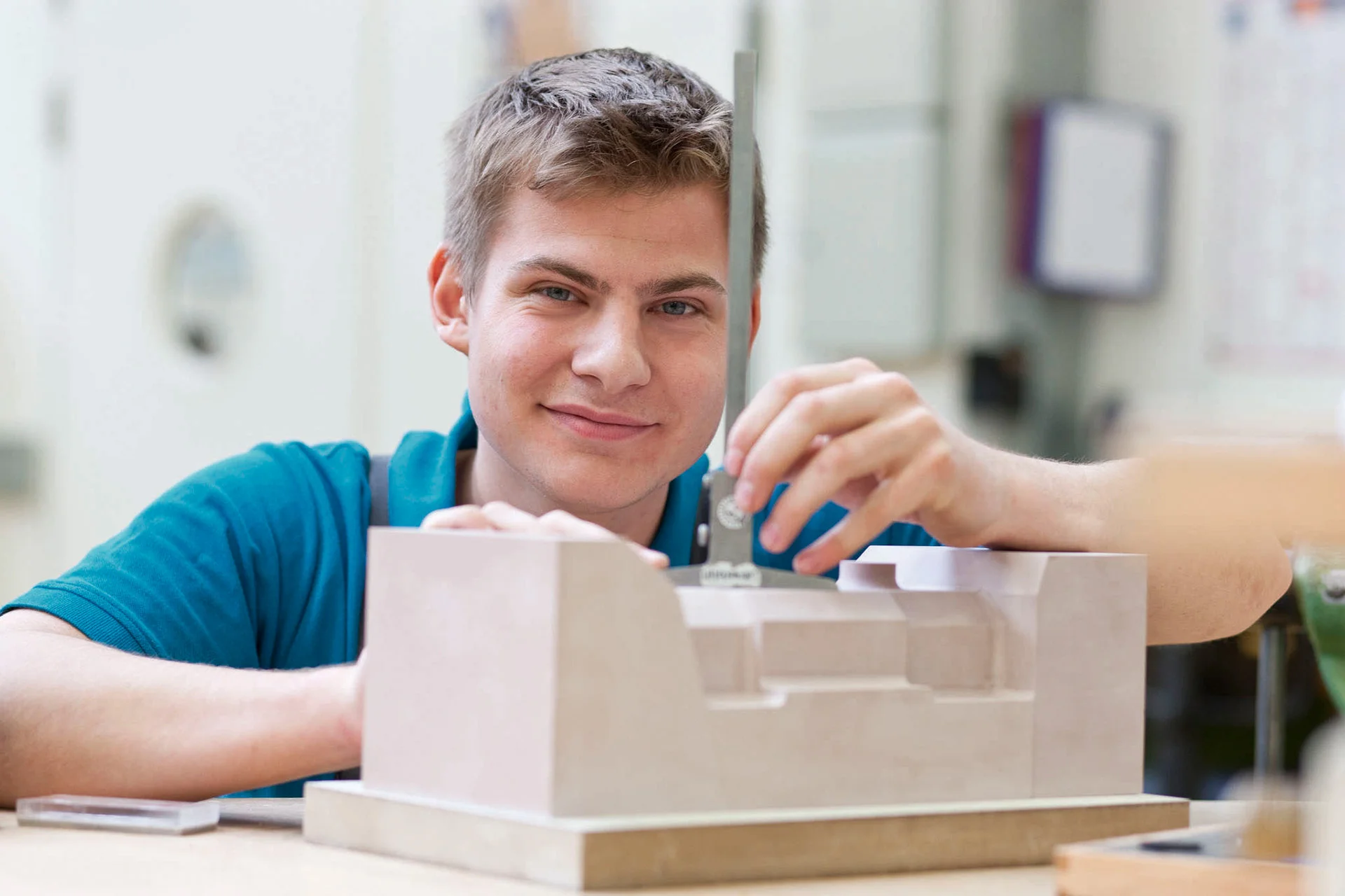 An apprentice at DRÄXLMAIER in Vilsbiburg smiles at the camera and holds a workpiece in his hand.