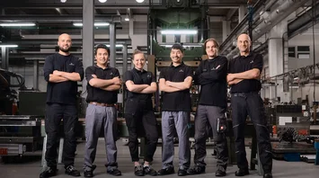 Six DRÄXLMAIER production employees stand in a row in a hall. They smile into the camera. 