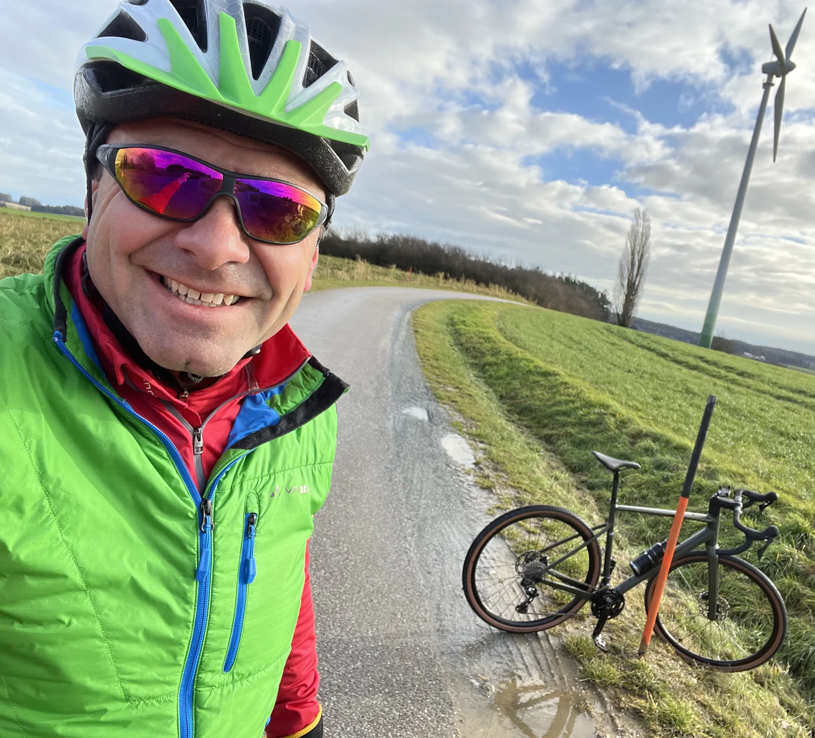 A man in cycling gear and helmet takes a selfie with his racing bike. He is outside on a small road.