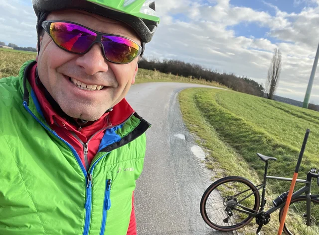 Ein Mann in Fahrradkleidung und Helm macht ein Selfie mit seinem Rennrad. Er befindet sich draußen auf einer kleinen Straße.