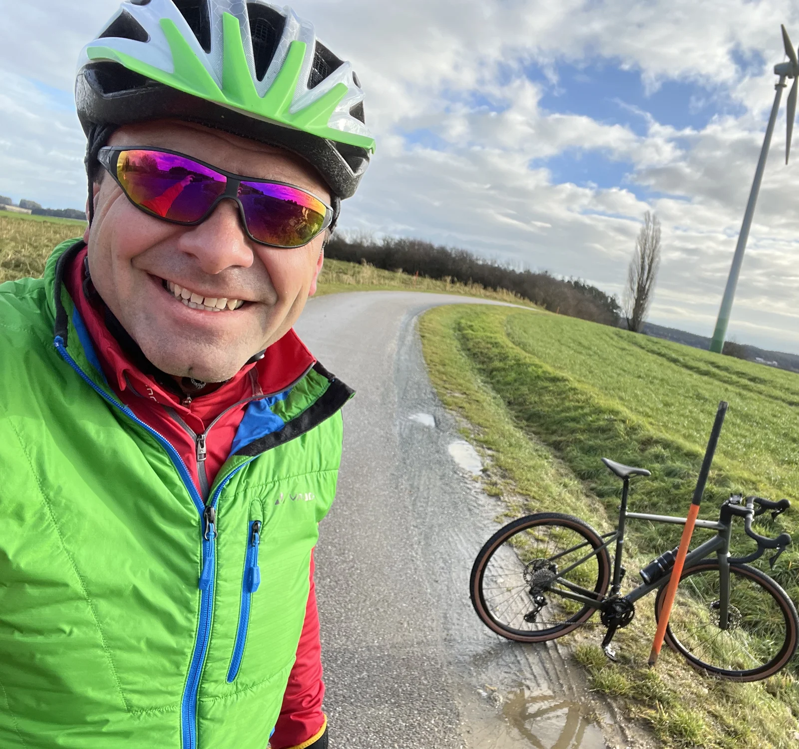 A man in cycling gear and helmet takes a selfie with his racing bike. He is outside on a small road.