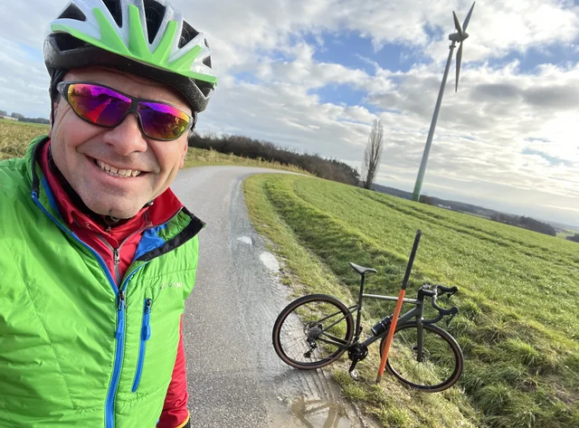 A man in cycling gear and helmet takes a selfie with his racing bike. He is outside on a small road.