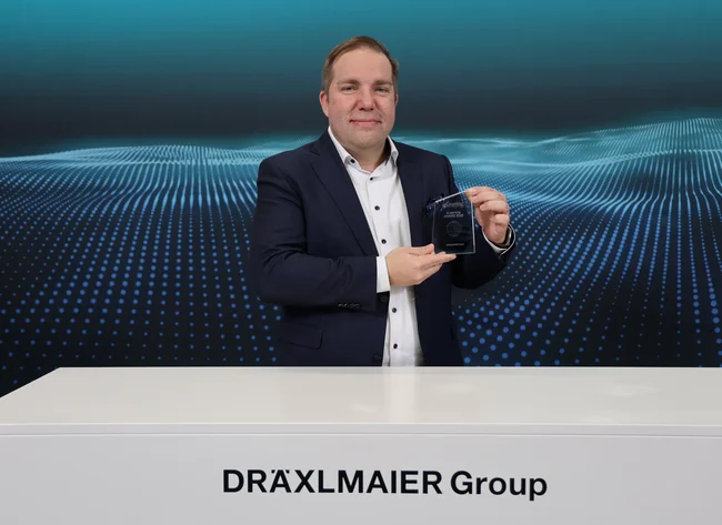 A man stands in front of a DRÄXLMAIER background and holds the eLearning Award in his hands.