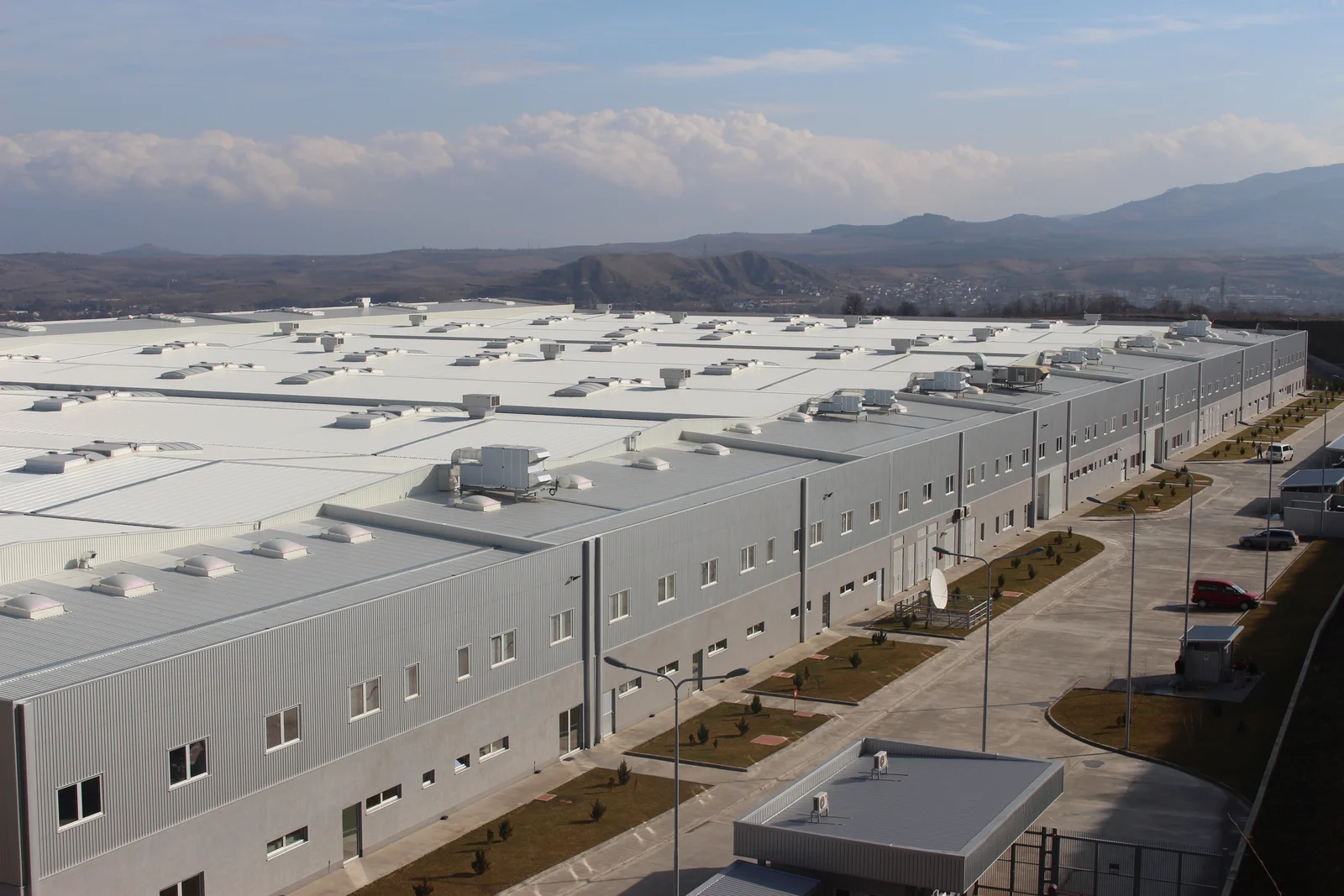 Große Industriehalle in Kavadarci, umgeben von einem Parkplatz und einer Landschaft mit Bergen im Hintergrund.