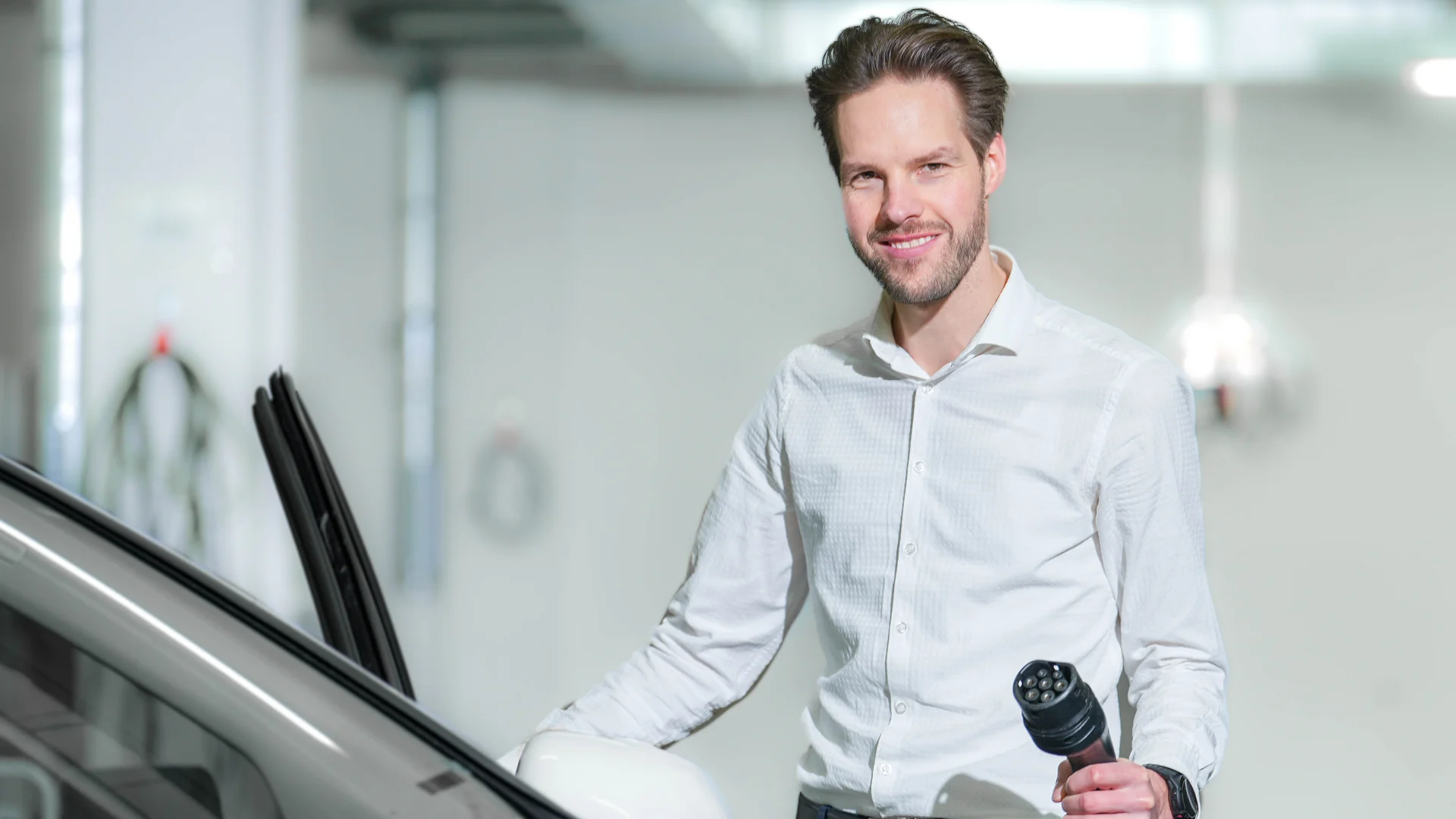 Man in a white formal shirt standing in front of an open car door with an electric charging plug in his hand.