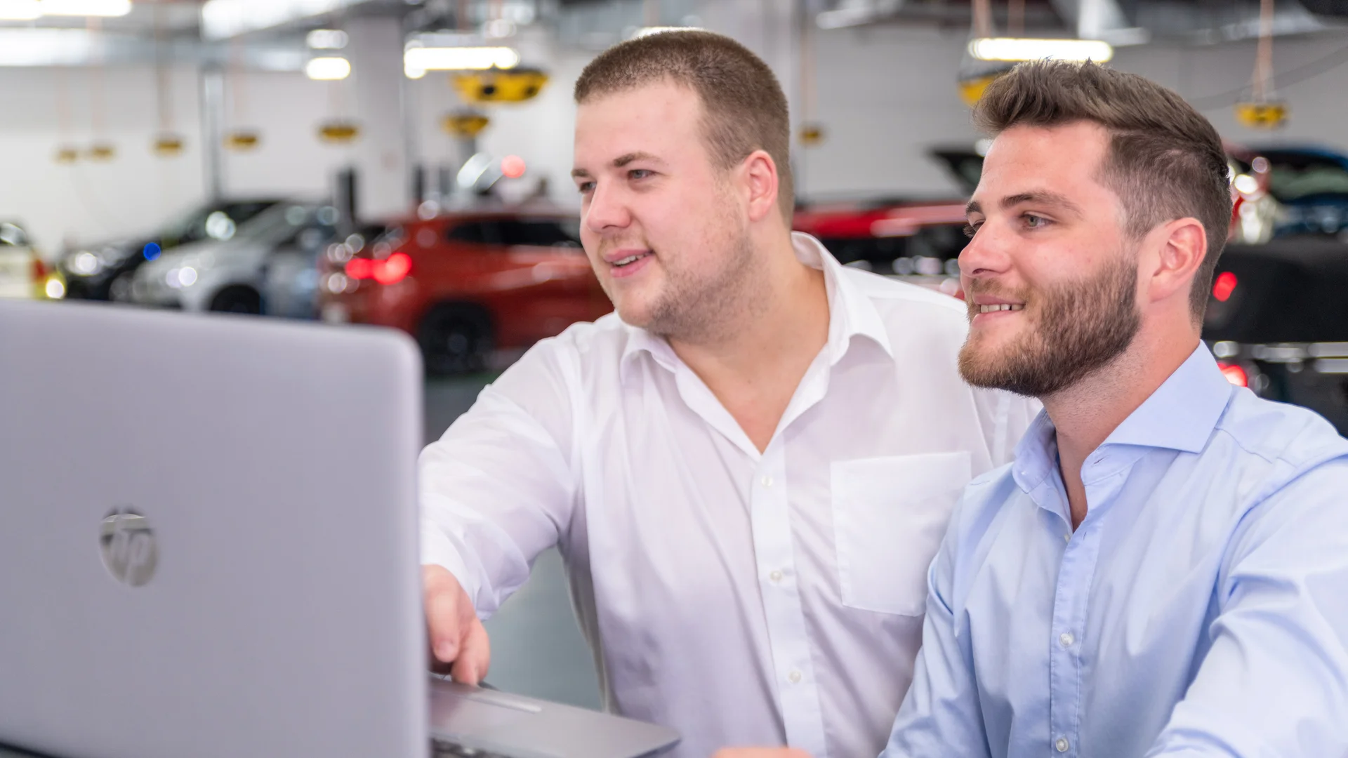 Two men in formal shirts are standing in a garage looking at a laptop.