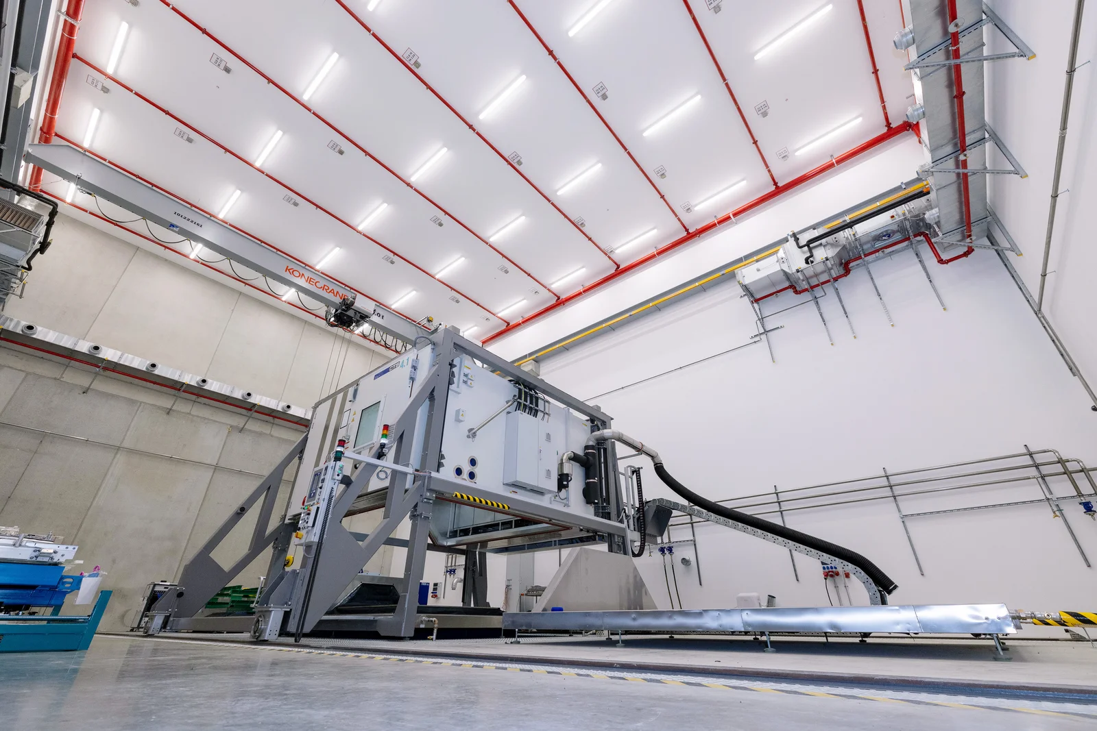 Large industrial machine in a spacious facility with high ceilings and various pipes and cables.