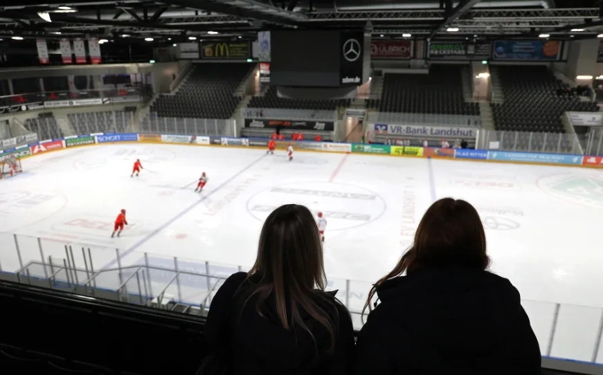 Eishockeyspiel in einer Halle mit Spielern in roten und weißen Trikots, zwei Zuschauern im Vordergrund und einem großen Bildschirm mit Mercedes-Benz-Logo.