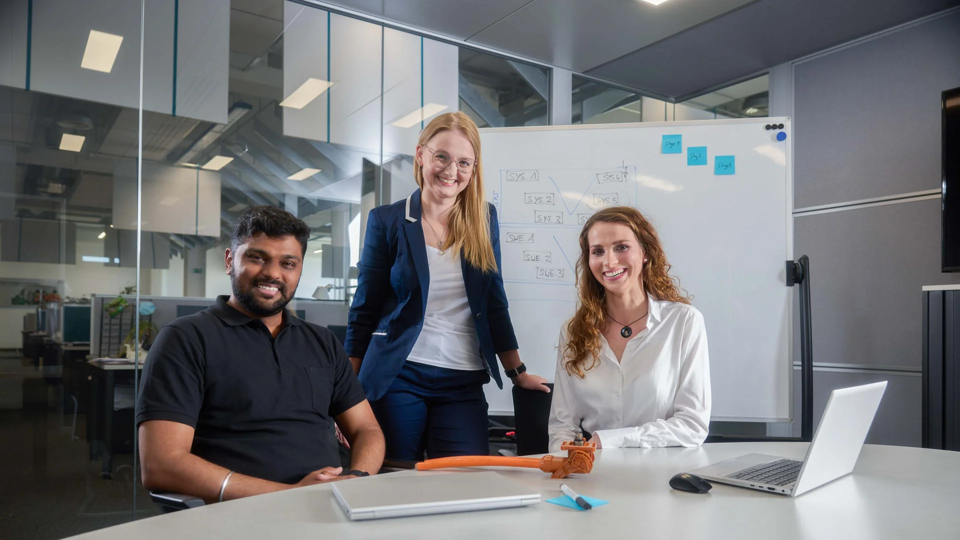 3 people are standing in front of a white board in a meeting room and smiling at the camera.