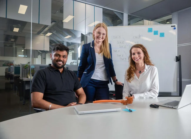 3 people are standing in front of a white board in a meeting room and smiling at the camera.
