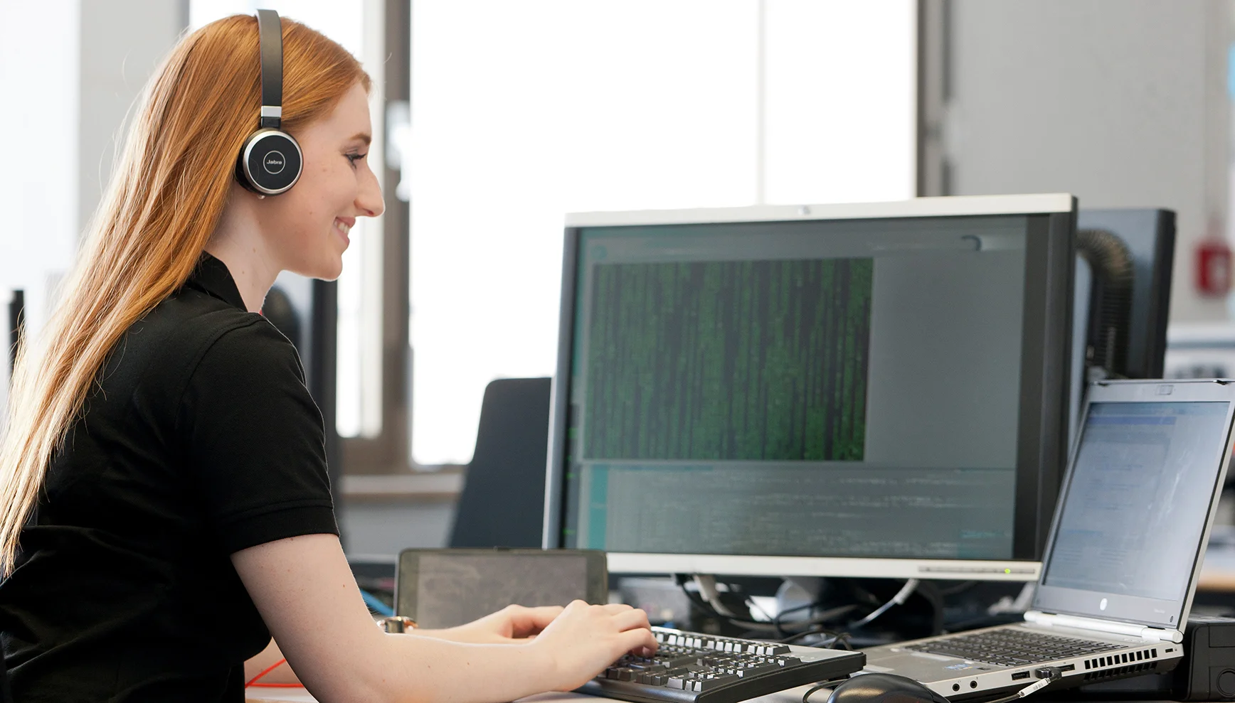 A trainee IT specialist for application development wears a headset and looks at the screen with a smile.