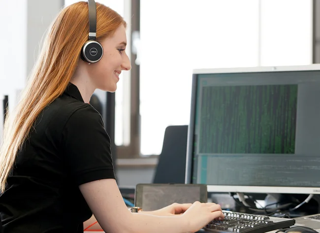 Eine Auszubildende zur Fachinformatikerin für Anwendungsentwicklung trägt ein Headset und schaut lächelnd auf den Bildschirm.