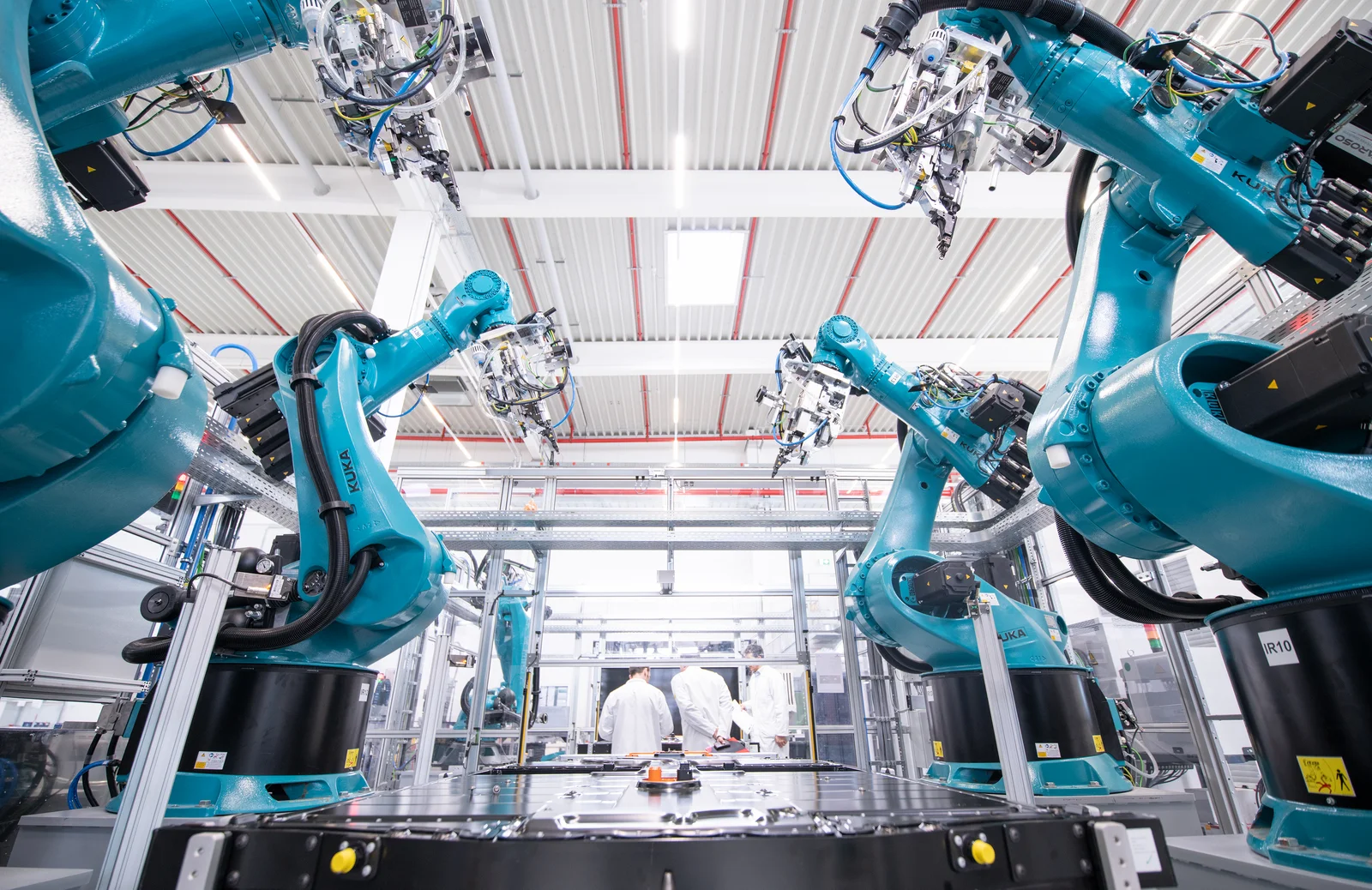 Mehrere blaue Industrieroboterarme in einer modernen Fabrikumgebung, drei Personen im Hintergrund.