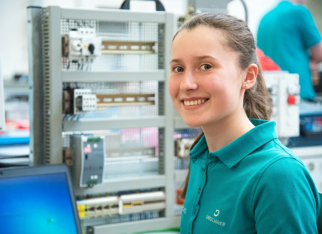 Eine Auszubildende bei DRÄXLMAIER in Vilsbiburg lächelt in die Kamera, während sie an einem technischen Projekt arbeitet.