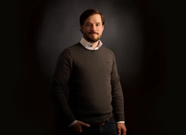 A man in front of a black background smiles into the camera with his arms crossed. 