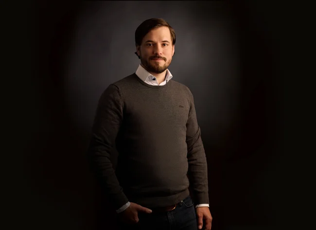 A man in front of a black background smiles into the camera with his arms crossed. 