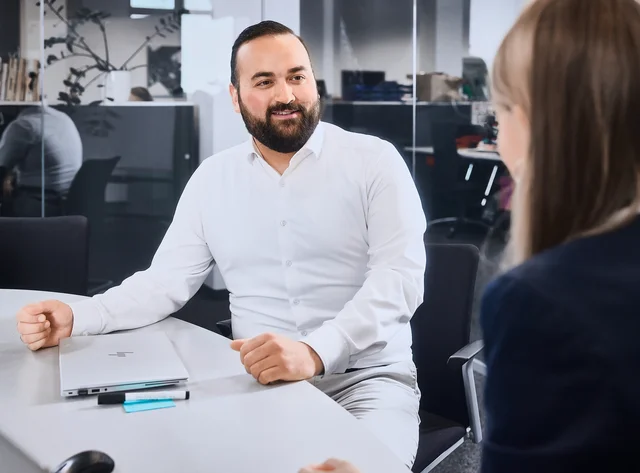 A man in a white formal shirt is sitting in a meeting room talking to the woman in front of him.